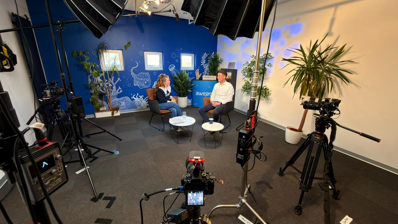 Moderatorin Melanie Donaldson im AWEOS-Hoodie im Gespräch mit einem Gast vor der blauen Studio-Wand in Solingen